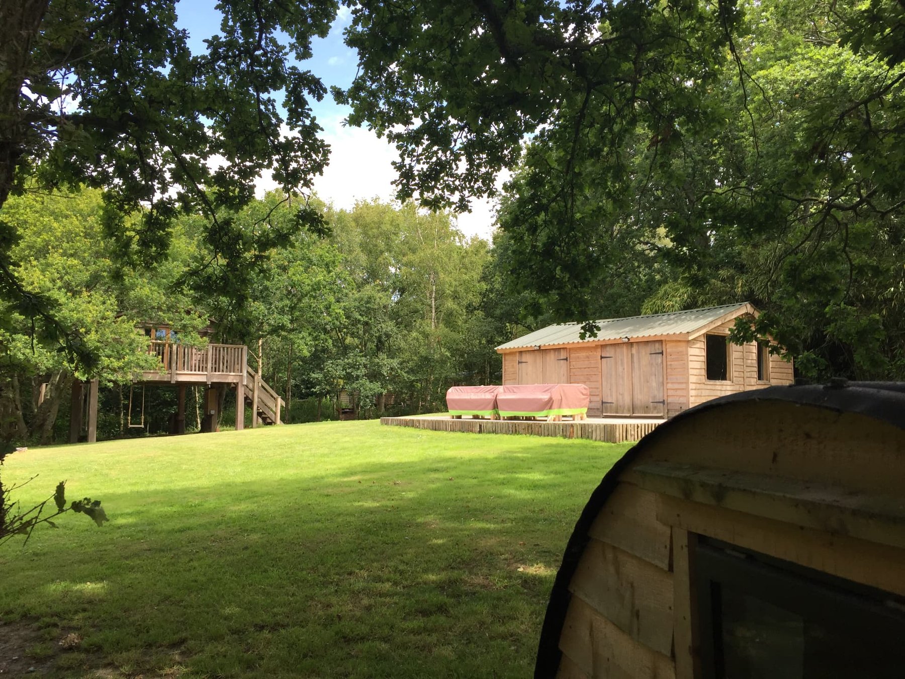 Into the Woods seen from the lawn — a treehouse on posts to the left, a timber building and deck with tables to the right, and a barrel-shaped pod in the foreground, framed by oak branches